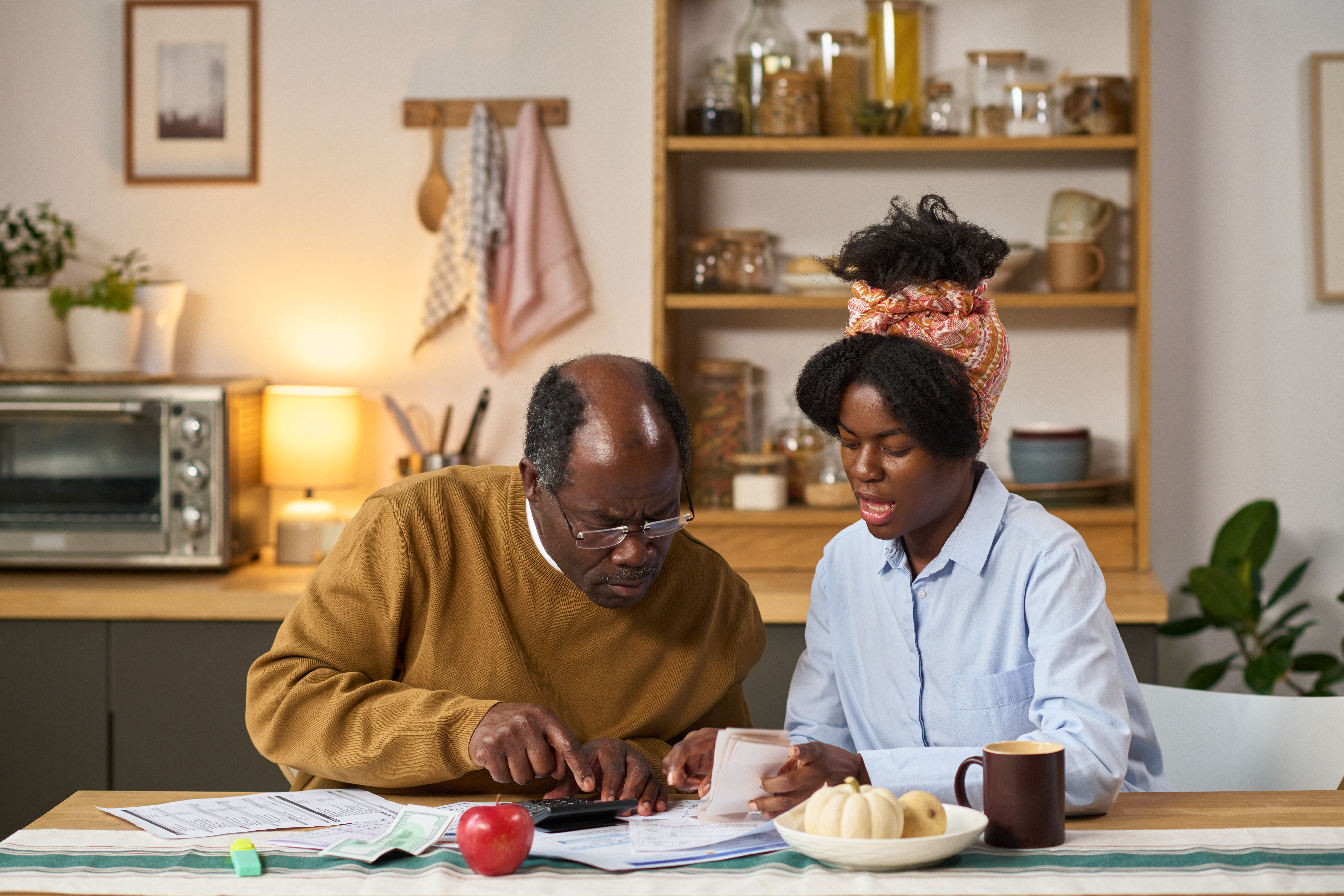 Middle aged Black man and young Black woman sitting at kitchen table reviewing financial documents and using calculator, focusing on paying taxes together, paperwork and receipts visible